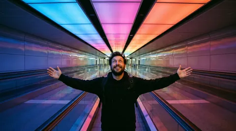 A man with arms spread wide in an empty airport walkway under a rainbow-lit ceiling